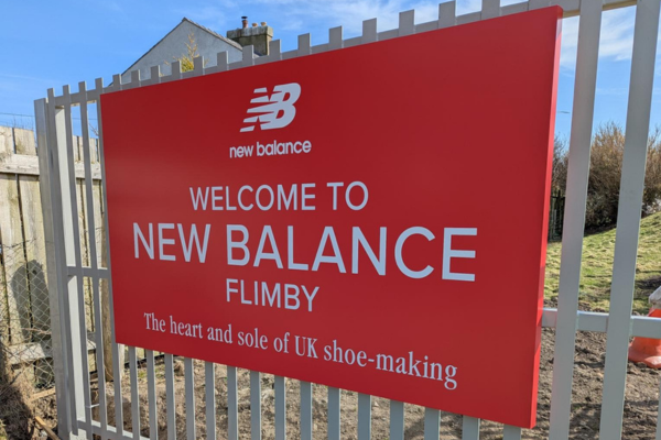 A red sign on the fence of an industrial yard which reads, "Welcome to New Balance Flimby - The Heart and Sole of UK Shoe-Making"