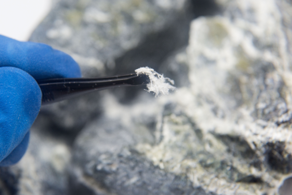 A close-up photo of a hand in a thick, blue glove holding a piece of asbestos with a pair of tweezers