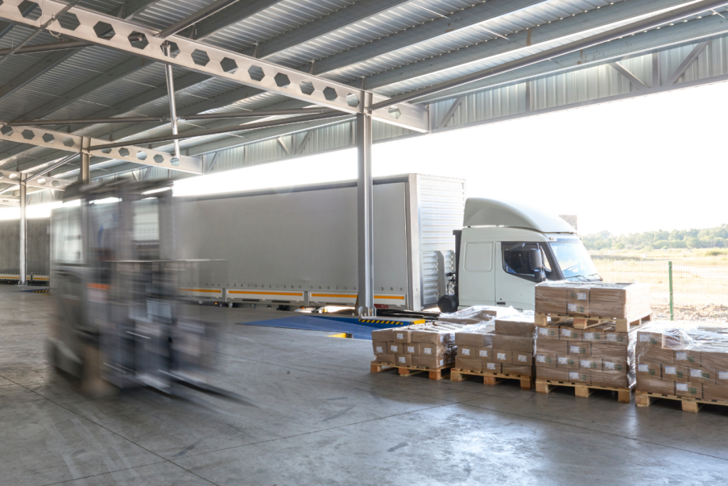 A lorry at a loading dock at a warehouse with pallets of goods in front of it and a blurred forklift in the foreground