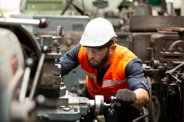 A man in a high-visibility jacket and wearing a white hard hat operating heavy machinery in a factory