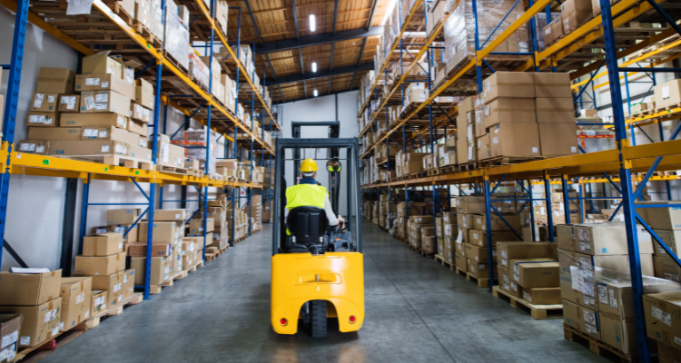 A person driving a forklift in a warehouse, which has shelves from floor to ceiling with cardboard boxes stacked on them.