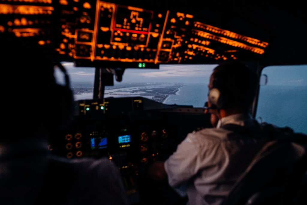 Pilot in the cockpit of a plane flying over seas during dark skies with lights showing for the buttons on the plane