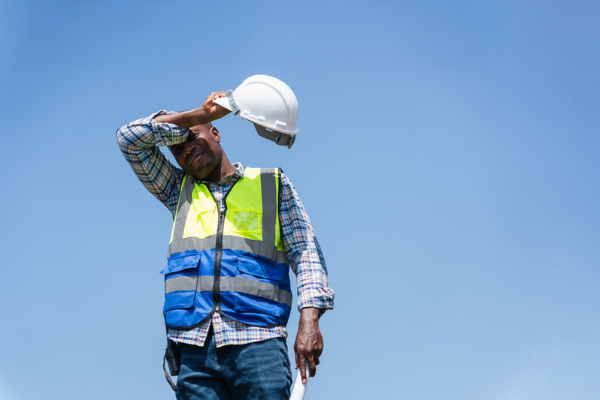 A photo of a construction worker stood against a bright blue sky, wiping sweat from his brow with his safety helmet in his hand